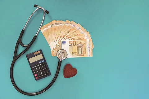 Stethoskop, Geldscheine, Taschenrechner und Stoffherz auf blauem Untergrund.