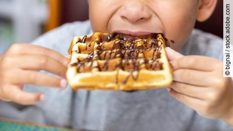 Kind beißt in eine belgische Waffel, die mit Schokoladensoße überzogen ist.