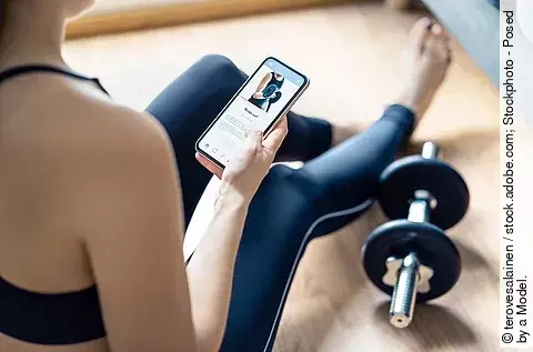 Frau sitzt auf einer Yoga-Matte und macht mit einer App Sport.