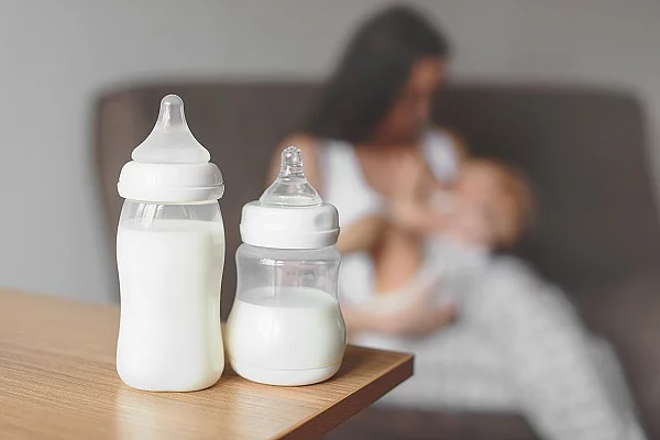 Zwei Flaschen mit Muttermilch stehen auf einem Tisch. Im Hintergrund hält eine Mutter ihr Baby.