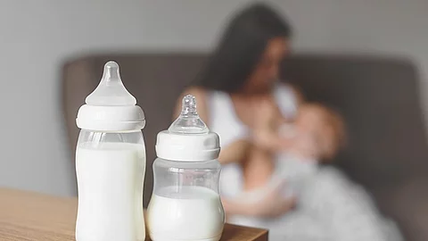 Zwei Flaschen mit Muttermilch stehen auf einem Tisch. Im Hintergrund hält eine Mutter ihr Baby. 