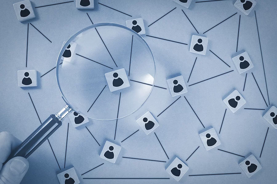 In einer schematischen Darstellungen sind Personen über ein Netzwerk verbunden. Eine Lupe vergrößert einen Ausschnitt des Netzwerks. In einer schematischen Darstellungen sind Personen über ein Netzwerk verbunden. Eine Lupe vergrößert einen Ausschnitt des Netzwerks.