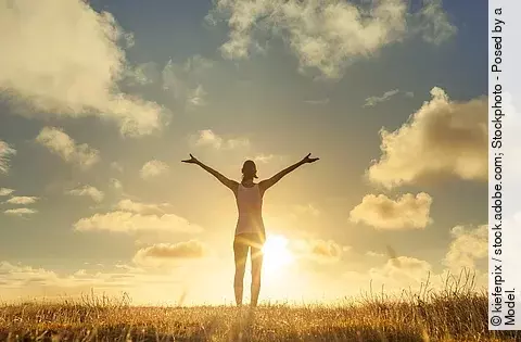 Frau steht bei Sonnenuntergang auf einem Feld und breitet die Arme aus.