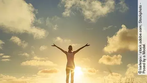 Frau steht bei Sonnenuntergang auf einem Feld und breitet die Arme aus.