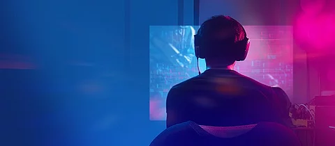 Mann trägt Kopfhörer und sitzt in einem dunklen Zimmer vor seinem PC.