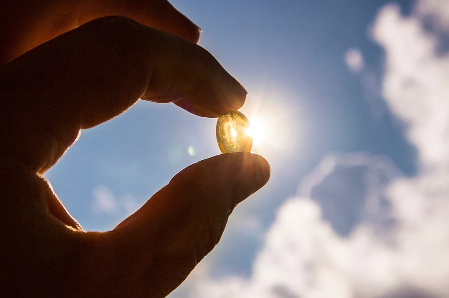 Eine Hand hält eine Vitamin-D-Kapsel vor sonnigem blauem Himmel