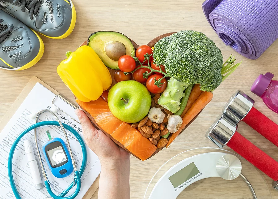 Symbolbild gesunder Lebensstil: Teller in Herzform mit Obst, Hanteln, Turnschuhe, Yogamatte