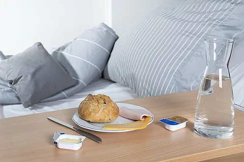Krankenhausbett, daneben Nachttisch mit Brötchen, Wurst, Käse, Margarine