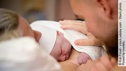 Ein Neugeborenes liegt auf der Brust der Mutter. Der Vater hat seine Hand auf den Rücken des Babys gelegt.