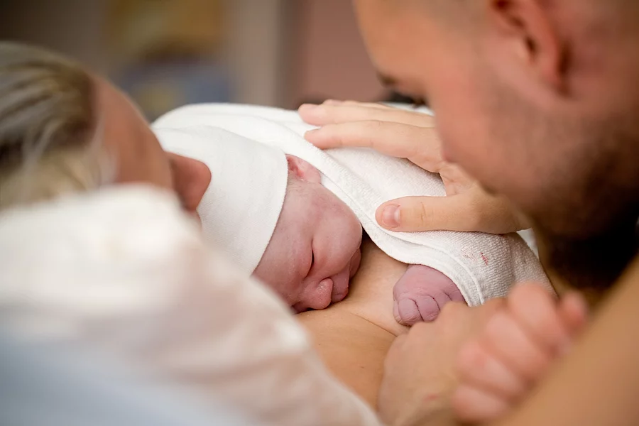 Ein Neugeborenes liegt auf der Brust der Mutter. Der Vater hat seine Hand auf den Rücken des Babys gelegt.