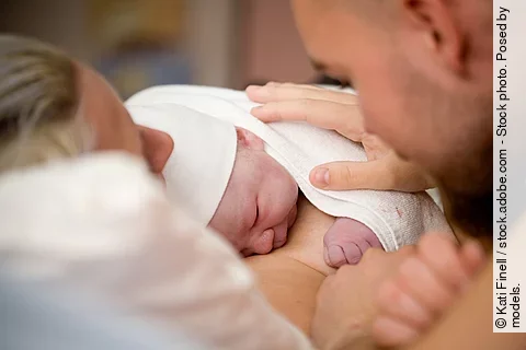 Ein Neugeborenes liegt auf der Brust der Mutter. Der Vater hat seine Hand auf den Rücken des Babys gelegt.