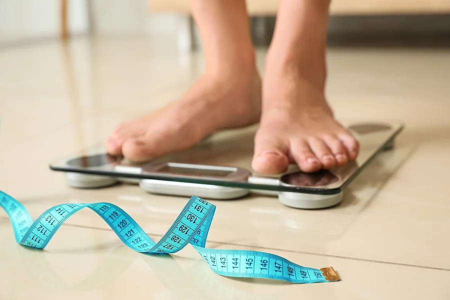 Tape in front of woman standing on floor scales indoors. Overweight problem Füße auf einer Personenwaage, davor liegt ein Maßband