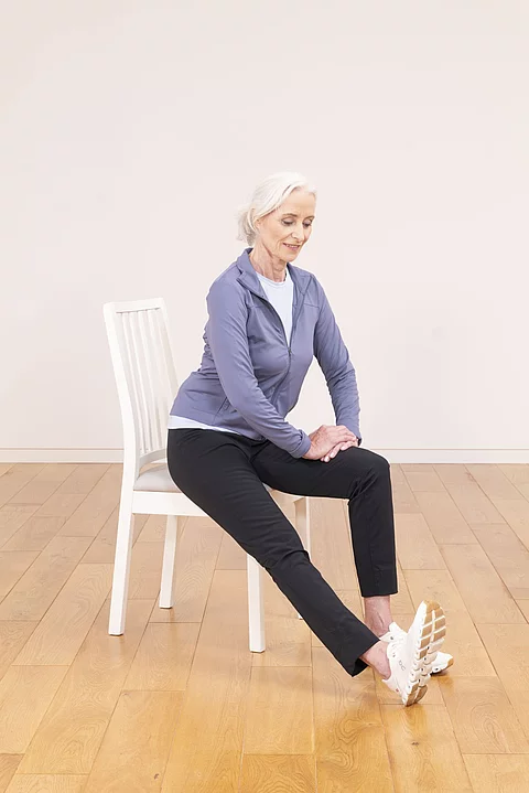 Seniorin in Trainingsanzug sitzt auf weißem Stuhl und macht Dehungsübungen