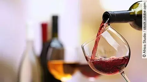 Es wird Rotwein in ein Glas gegossen. Im Hintergrund befinden sich weitere Weinflaschen und Gläser. 