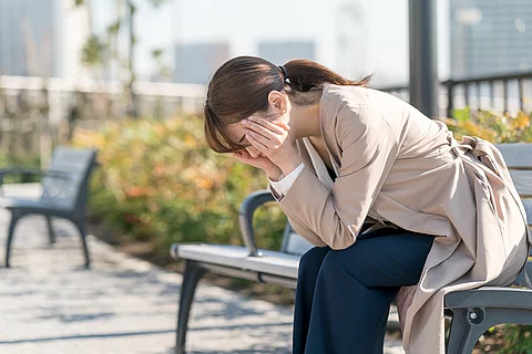 Erschöpfte Frau sitzt auf einer Parkbank, nach vorn gebeugt mit Kopf in den Händen