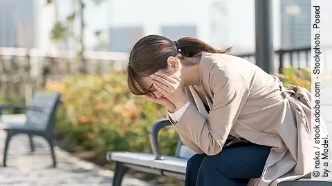 Erschöpfte Frau sitzt auf einer Parkbank, nach vorn gebeugt mit Kopf in den Händen