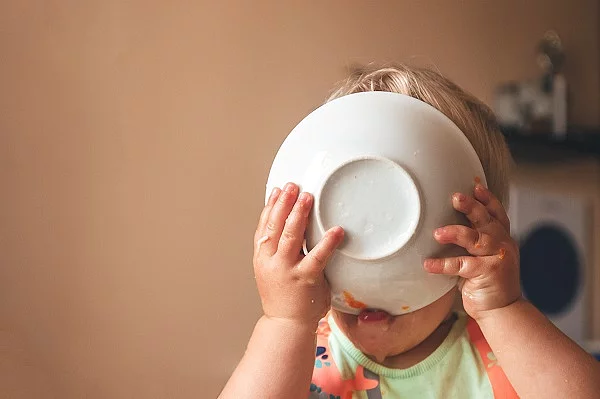 Kleinkind, das hungrig den Teller vor das Gesicht hält und ableckt.