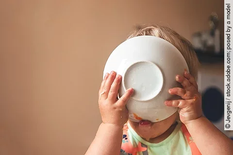 Kleinkind, das hungrig den Teller vor das Gesicht hält und ableckt.