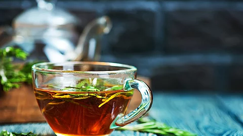 Glas mit brauner Flüssigkeit und einzelnen Pflanzenteilen darin auf einem blau gestrichenen Holztisch mit einer verschwommenen Teekanne im Hintergrund.