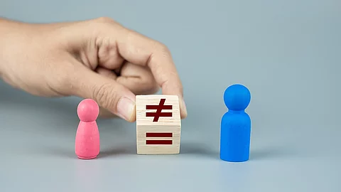 Pinke und blaue Spielfigur zwischen denen ein Holzwürfel, mit dem Ungleich-Zeichen, von einer Hand festgehalten wird. 