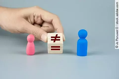 Pinke und blaue Spielfigur zwischen denen ein Holzwürfel, mit dem Ungleich-Zeichen, von einer Hand festgehalten wird. 