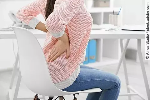 Eine Frau sitzt an einem Schreibtisch und hat beide Hände auf den unteren Rücken gestützt. 
