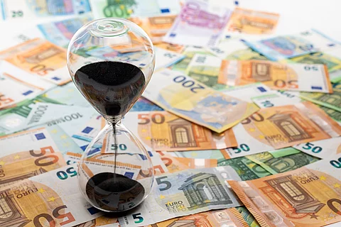 Geldscheine verteilt auf dem Boden. Eine Sanduhr steht auf den Scheinen.