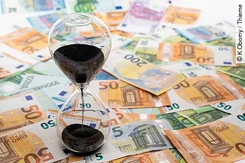 Geldscheine verteilt auf dem Boden. Eine Sanduhr steht auf den Scheinen.