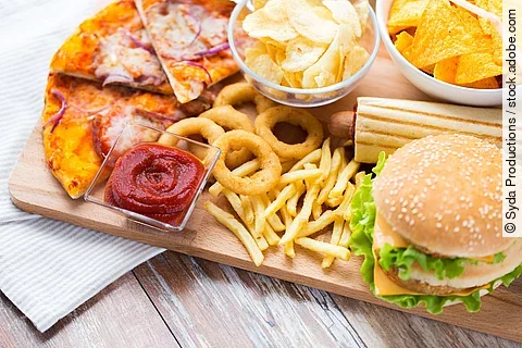 Hochverarbeitete Lebensmittel: Cheeseburger, Pommes, Chips, Ketchup, Pizza auf einem Holzbrett