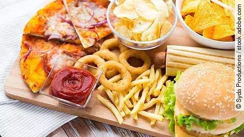 Hochverarbeitete Lebensmittel: Cheeseburger, Pommes, Chips, Ketchup, Pizza auf einem Holzbrett