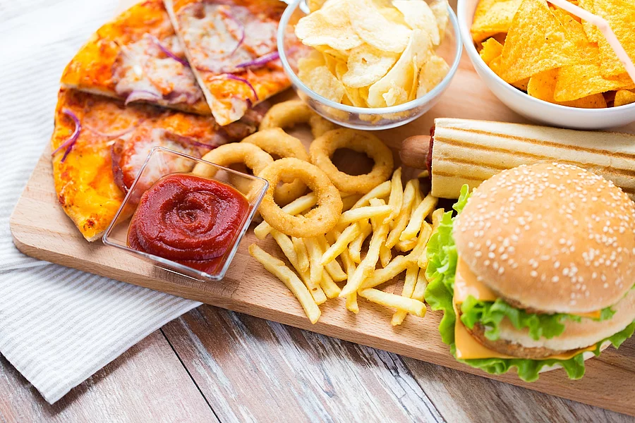Hochverarbeitete Lebensmittel: Cheeseburger, Pommes, Chips, Ketchup, Pizza auf einem Holzbrett