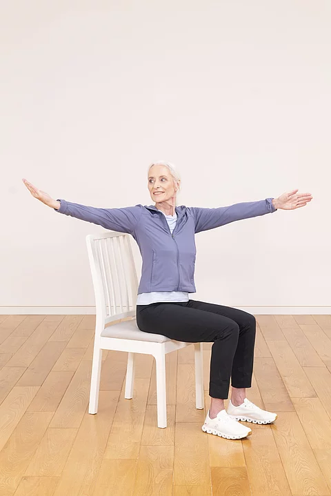Seniorin sitzt mit ausgebreiteten Armen auf weißem Stuhl 