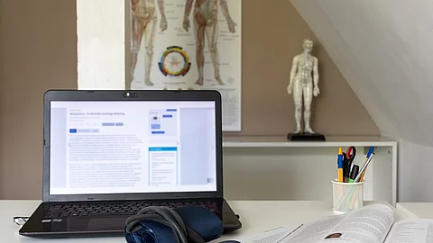 Symbolbild Integrative Medizin: Schreibtisch mit Laptop, Stethoskop, im Hintergrund Plakat mit TCM-Meridianen 