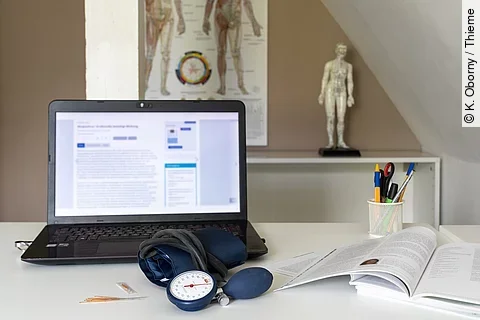 Symbolbild Integrative Medizin: Schreibtisch mit Laptop, Stethoskop, im Hintergrund Plakat mit TCM-Meridianen 
