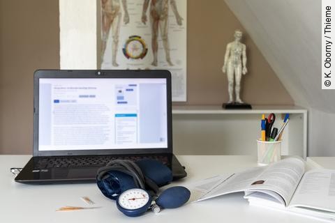 Symbolbild Integrative Medizin: Schreibtisch mit Laptop, Stethoskop, im Hintergrund Plakat mit TCM-Meridianen 