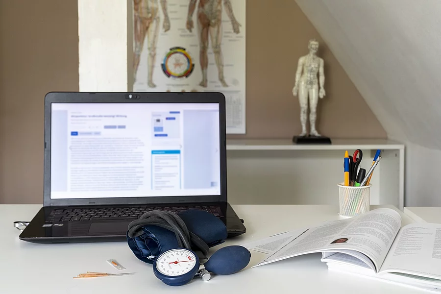 Symbolbild Integrative Medizin: Schreibtisch mit Laptop, Stethoskop, im Hintergrund Plakat mit TCM-Meridianen 