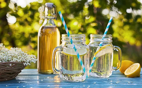 Flasche mit heller Flüssigkeit sowie zwei Gläser ebenfalls mit heller Flüssigkeit gefüllt sowie je einer Scheibe Zitrone und einem Trinkhalm auf einem blauen Tisch vor verschwommenem Hintergrund.