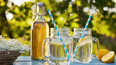 Flasche mit heller Flüssigkeit sowie zwei Gläser ebenfalls mit heller Flüssigkeit gefüllt sowie je einer Scheibe Zitrone und einem Trinkhalm auf einem blauen Tisch vor verschwommenem Hintergrund.