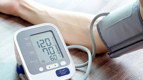 Blutdruckmessgerät im Vordergrund. Im Hintergrund befindet sich der Arm einer Person, die ihren Blutdruck misst.