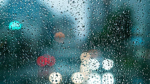 Trüber Herbsttag, durch eine Fensterscheibe mit Regentropfen sieht man Autolichter auf einer Straße und im Hintergrund verschwommene Häuserkonturen