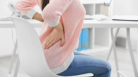 Frau sitzt auf einem Stuhl und hält sich den schmerzenden Rücken Frau sitzt auf einem Stuhl und hält sich den schmerzenden Rücken