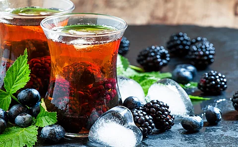 Zwei Gläser mit Eistee mit ganzen Brombeeren im Glas. Um die Gläser herum sind Brombeeren, Brombeerblätter, Heidelbeeren und Eiswürfel als Dekoration verstreut. 