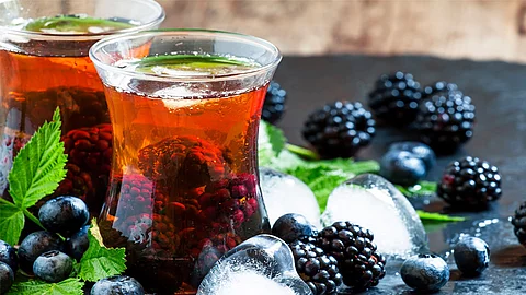 Zwei Gläser mit Eistee mit ganzen Brombeeren im Glas. Um die Gläser herum sind Brombeeren, Brombeerblätter, Heidelbeeren und Eiswürfel als Dekoration verstreut.