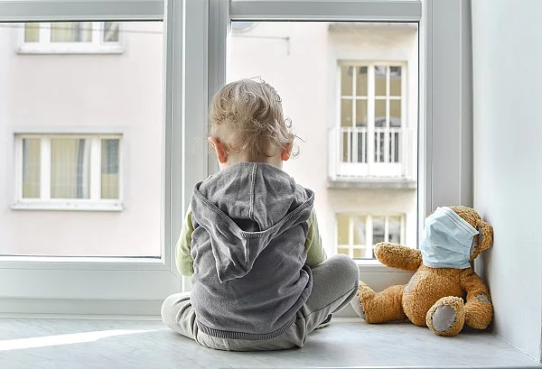 Kind sitzt alleine vor dem Fenster und blickt hinaus. Rechts daneben ist ein Teddybär, der eine Maske trägt. 