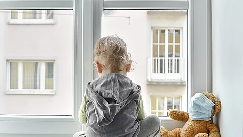 Kind sitzt alleine vor dem Fenster und blickt hinaus. Rechts daneben ist ein Teddybär, der eine Maske trägt. 