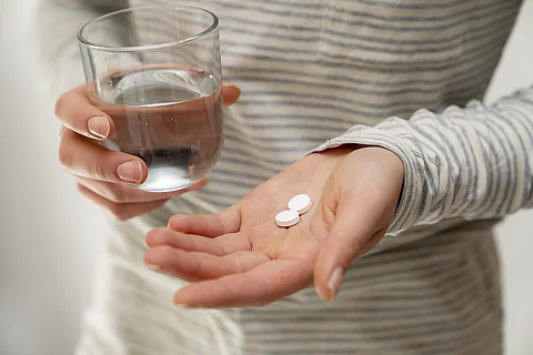 Eine Person hält in der flachen Hand zwei weiße Tabletten und in der anderen Hand ein Glas Wasser.