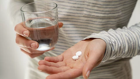 Eine Person hält in der flachen Hand zwei weiße Tabletten und in der anderen Hand ein Glas Wasser.