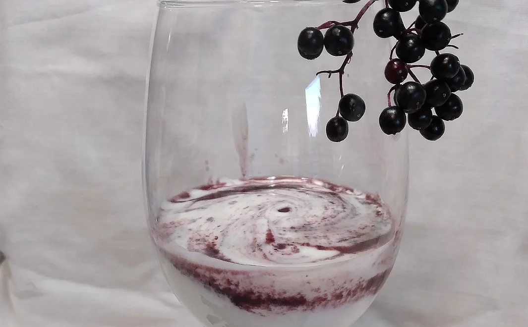 Ein Glas gefüllt mit weißem Sahnequark und Holunderbeerenmarmelade zu Schlieren verrührt, dekoriert mit schwarzen Holunderbeeren.