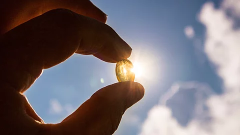 Hand hält Vitamin D-Kapsel vor sonnigen blauen Himmel