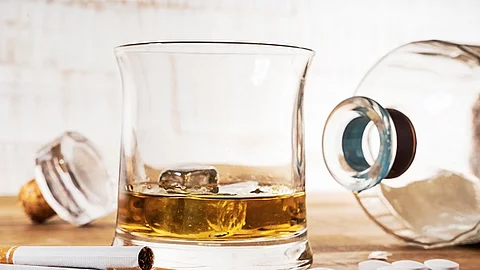 Zigaretten, Tabletten, eine liegende Glasflasche und ein Glas mit Whiskey sind auf einem Holztisch ausgebreitet.